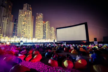L'image montre une scène de cinéma en plein air sur une plage, animée pendant la nuit. Un grand écran est installé, et des groupes de personnes s'asseyent sur des poufs colorés, principalement de couleur rouge. En arrière-plan, on peut voir des gratte-ciels illuminés, créant une atmosphère vibrante et moderne. L'éclairage doux et les ombres ajoutent à l'ambiance festive du lieu.