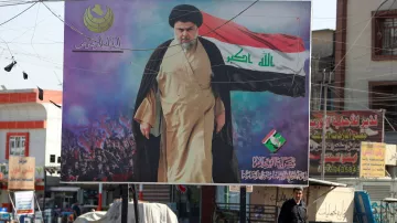 L'image montre une grande affiches publicitaire dans un espace urbain. Au centre, un homme en costume traditionnel, probablement un religieux, est représenté avec un drapeau. Le fond de l'affiche semble comporter une foule, ce qui suggère un événement ou un rassemblement. Une personne marche devant l'affiche, ajoutant une dimension humaine à la scène. Les couleurs dominantes sont le noir, le rouge et le vert, qui sont souvent symboliques dans le contexte politique.