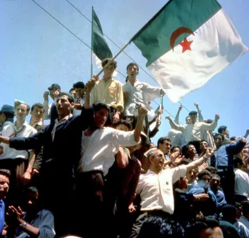 L'image montre une foule rassemblée, levant des bras et brandissant des drapeaux, dont celui de l'Algérie. Les personnes semblent joyeuses et engagées, célébrant un moment fort. L'atmosphère pourrait évoquer une célébration de l'indépendance ou un événement historique significatif pour le pays. Les visages sont animés et l'énergie collective est palpable.