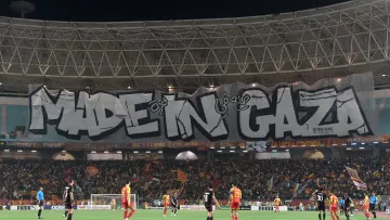 L'image montre un stade de football avec une grande banderole affichée. La banderole est ornée du texte "MADE IN GAZA" écrit en lettres stylisées et imposantes. Le stade est rempli de supporters, et l'atmosphère semble animée. On peut voir des joueurs sur le terrain, ce qui indique qu'un match est en cours. L'ambiance est festive et engagée.
