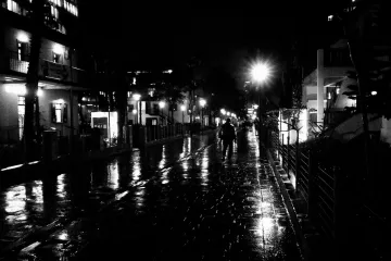 L'image montre une rue mouillée de nuit, éclairée par des lampadaires. Le sol reflète la lumière, créant un effet brillant. On peut voir quelques personnes marchant sur le trottoir, tandis que des bâtiments se dressent de chaque côté, illuminés par des lumières. L'atmosphère semble calme et sereine, typique d'une soirée de pluie.