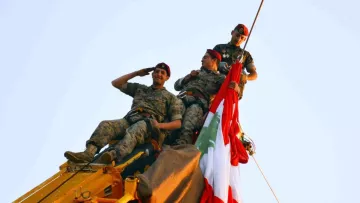 L'image montre trois soldats en uniforme, perchés en haut d'une grue. Ils sont en train de saluer tout en tenant un grand drapeau, probablement celui du Liban. Les soldats affichent un air déterminé et solennel, et l'ambiance semble festive, suggérant une célébration ou un événement important. Le ciel est clair, ce qui met en valeur la scène.