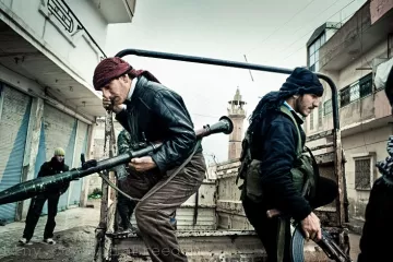 L'image montre des individus armés, apparemment dans un environnement urbain. Deux hommes sont en mouvement, l'un portant une arme lourde et l'autre un fusil. Ils semblent faire partie d'un groupe en action, probablement dans un contexte de conflit. L'arrière-plan montre des bâtiments et une ambiance sombre, qui renforcent le sentiment de tension. L'atmosphère est chargée et suggère un moment urgent.
