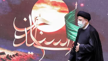 در این تصویر، یک مرد در لباس رسمی و با عمامه مشکی در حال راه رفتن است. او ماسک به صورت دارد و پشت او تصاویری از یک پس‌زمینه هنری با رنگ‌های گرم و نمادهایی مربوط به تاریخ یا فرهنگ دیده می‌شود، از جمله پرچم سبز. متن خوشنویسی شده‌ای در تصویر وجود دارد که به زبان فارسی نوشته شده است. این تصویر ممکن است به یک مراسم مذهبی یا ملی مرتبط باشد.