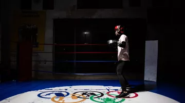 L'immagine mostra un giovane pugile che indossa i guantoni e un casco protettivo, mentre si allena in un ring da boxe. Lo sfondo è parzialmente illuminato, mettendo in risalto il pugile in azione. Sotto di lui, si trova un logo con i simboli olimpici e la scritta "ELBARRIO", che suggerisce un ambiente dedicato allo sport e alla formazione. La scena ha un'atmosfera di concentrazione e determinazione.