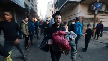 L'immagine mostra un gruppo di persone che si muovono in una strada affollata. Un uomo in primo piano, visibilmente agitato, sta correndo e tiene in braccio dei vestiti. Gli altri presenti nella scena sembrano essere coinvolti in una situazione di emergenza o caos, contribuendo a creare un'atmosfera di urgenza e tensione. Gli edifici circostanti appaiono deteriorati, suggerendo un contesto di conflitto o crisi.