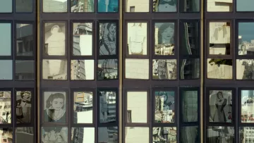 L'image montre une façade de bâtiment avec de grandes fenêtres. À travers ces fenêtres, on peut voir des reflets de la ville environnante ainsi que des illustrations ou des portraits de personnes qui semblent être intégrés dans les carreaux. Les illustrations sont stylisées et en noir et blanc, ajoutant un contraste intéressant avec les reflets colorés des bâtiments environnants. Cette composition crée une atmosphère visuelle dynamique et intrigante.