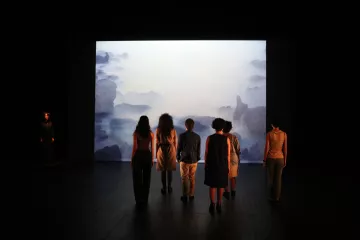 L'image montre une scène de performance artistique. On y voit plusieurs personnes de dos, regardant un grand écran qui projette une image évoquant un paysage marin brumeux. En arrière-plan, on aperçoit une silhouette d'une personne habillée en noir, se tenant à gauche de l'image. Les autres participants, vêtus de différentes tenues, semblent absorbés par la projection. L'éclairage est tamisé, créant une ambiance contemplative.