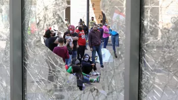 L'image montre un groupe de personnes rassemblées à l'extérieur, vues à travers un verre brisé. On peut voir une ambiance de tension, probablement liée à une manifestation ou un rassemblement. Certains individus portent des masques, ce qui peut indiquer une préoccupation pour la santé ou une mesure de protection dans un contexte de protestation. L'arrière-plan contient des éléments de graffiti, suggérant une scène urbaine dynamique.