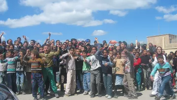 تظهر في الصورة مجموعة كبيرة من الأطفال والشباب يتجمعون في مكان واحد، يبدو أنهم متحمسون ويقومون بالتفاعل مع شيء ما، ربما يجتمعون لمتابعة حدث أو نشاط معين. السماء في الخلفية زرقاء مع بعض السحب، والمكان يبدو كأنه في منطقة حضرية. يرتدي الأطفال ملابس متنوعة، وتعكس تعبيراتهم الحماس والإثارة.