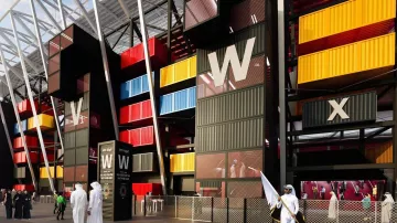 The image depicts a modern architectural structure that resembles a stadium or a large public venue. The building features a striking façade made up of brightly colored shipping containers in red, yellow, and blue, arranged in a visually dynamic way. There are tall, angular elements with a white roof framework above. In the foreground, a group of people dressed in traditional attire can be seen, indicating a cultural context. Overall, the design conveys a sense of contemporary style and innovative use of space.