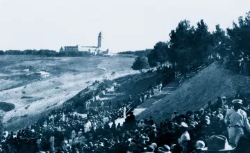 A large crowd in historical clothing gathers on a hillside, with a building in the distance.