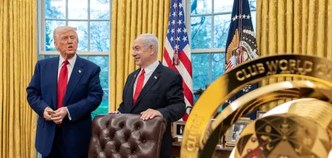 Two men in suits stand in an ornate office with gold curtains and flags, one holding a small object.