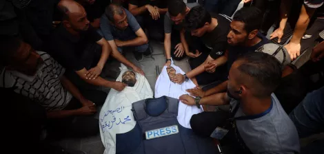 A group of people mourns over a deceased individual in a white shroud, surrounded by a press vest.