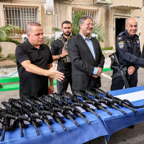 Men in uniform gather around rifles on a table, exchanging greetings and smiles.