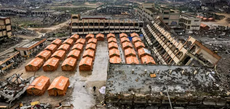 L'immagine mostra un'area devastata, con edifici in rovina sullo sfondo e tende arancioni disposte ordinatamente in primo piano. Il terreno sembra sporco e disordinato, evidenziando una situazione di emergenza o di sfollamento.
