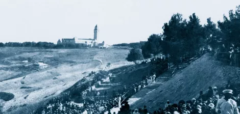 A large crowd in historical clothing gathers on a hillside, with a building in the distance.