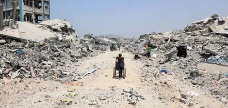 Una strada polverosa tra macerie di edifici distrutti, con una persona in sedia a rotelle.