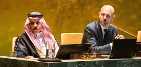 Two men at a conference table, one in traditional attire, discussing on a stage with microphones.