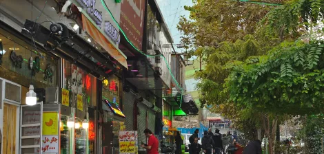خیابان شلوغ با فروشگاه‌ها و تبلیغات رنگارنگ، مردمی در حال عبور و درختان سبز در کناره‌ها.