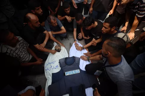 A group of people mourns over a deceased individual in a white shroud, surrounded by a press vest.