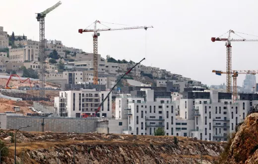 L'image montre un paysage urbain en construction. On peut voir plusieurs grues de construction sur des bâtiments en cours d'érection. Les structures, encore inachevées, présentent des façades en béton. À l'arrière-plan, on aperçoit des collines et d'autres constructions, indiquant un développement résidentiel dans la région. Le ciel semble brumeux, ce qui donne une ambiance un peu sombre à la scène.