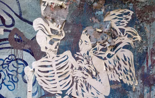 L'image montre une œuvre de street art sur un mur usé. On peut y voir un squelette et une figure féminine aux ailes, tous deux découpés dans un matériau qui semble être du papier ou du carton. Le fond est composé de textures et de couleurs dégradées, avec des traces d'usure et de peeling. L'ensemble dégage une atmosphère mystérieuse et à la fois poétique, mettant en avant les thèmes de la vie et de la mort.