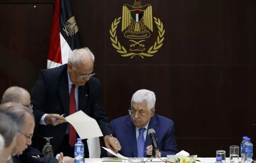 L'image montre une réunion, probablement politique, avec plusieurs hommes assis à une table. Au premier plan, un homme, probablement un leader ou un officiel, est assis regardant des documents. Un autre homme, debout à ses côtés, semble lui présenter des papiers ou des informations. En arrière-plan, on peut voir un drapeau, ainsi qu'un emblème qui semble représenter une organisation ou un gouvernement. L'ambiance générale de la scène est sérieuse et attentive, suggérant une discussion importante.