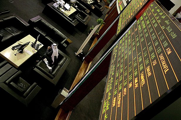 The image depicts a financial trading environment, likely within a stock exchange. In the foreground, there is a large electronic display board showing various financial data, such as stock prices and indices, with green and red text indicating changes in value. Below the board, there are several black leather seating areas where traders or visitors might be sitting. One individual, dressed in traditional attire, appears to be engaged with a computer or taking notes. The overall atmosphere suggests a busy and professional financial setting.