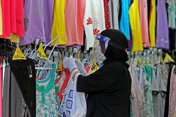 The image depicts a person wearing a black hijab and a face shield, shopping for clothing in a store. They are holding a white shopping bag and carefully examining different tops displayed on hangers. The store features a colorful array of clothing items, including various shades of purple, yellow, and floral patterns, hanging on racks in the background. The environment suggests a retail space focused on apparel, likely catering to a diverse range of styles.