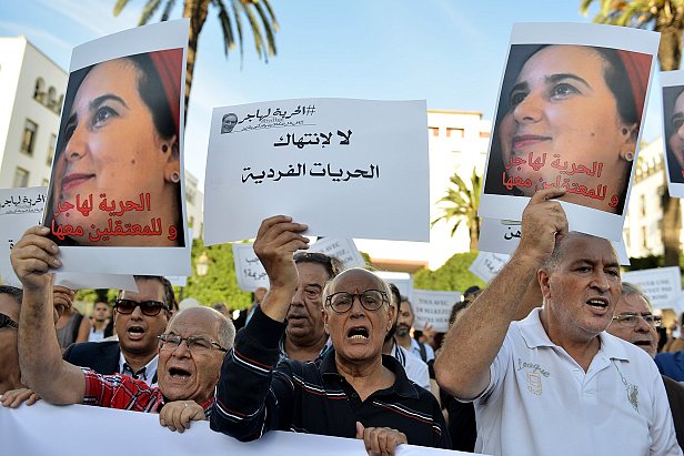 تظهر في الصورة مجموعة من الناس يتظاهرون في مكان عام، حاملين لافتات مكتوبة. يبدو أنهم يعبرون عن مطالب تتعلق بالحرية الفردية وحقوق المعتقلين. المشهد يدل على حماس المشاركين، حيث يعبر بعضهم عن استيائهم بصوت عالٍ. في الخلفية، يمكن رؤية بعض النخيل والسماء التي تحتوي على بعض السحب، مما يدل على أن المظاهرة جرت في يوم مشمس.